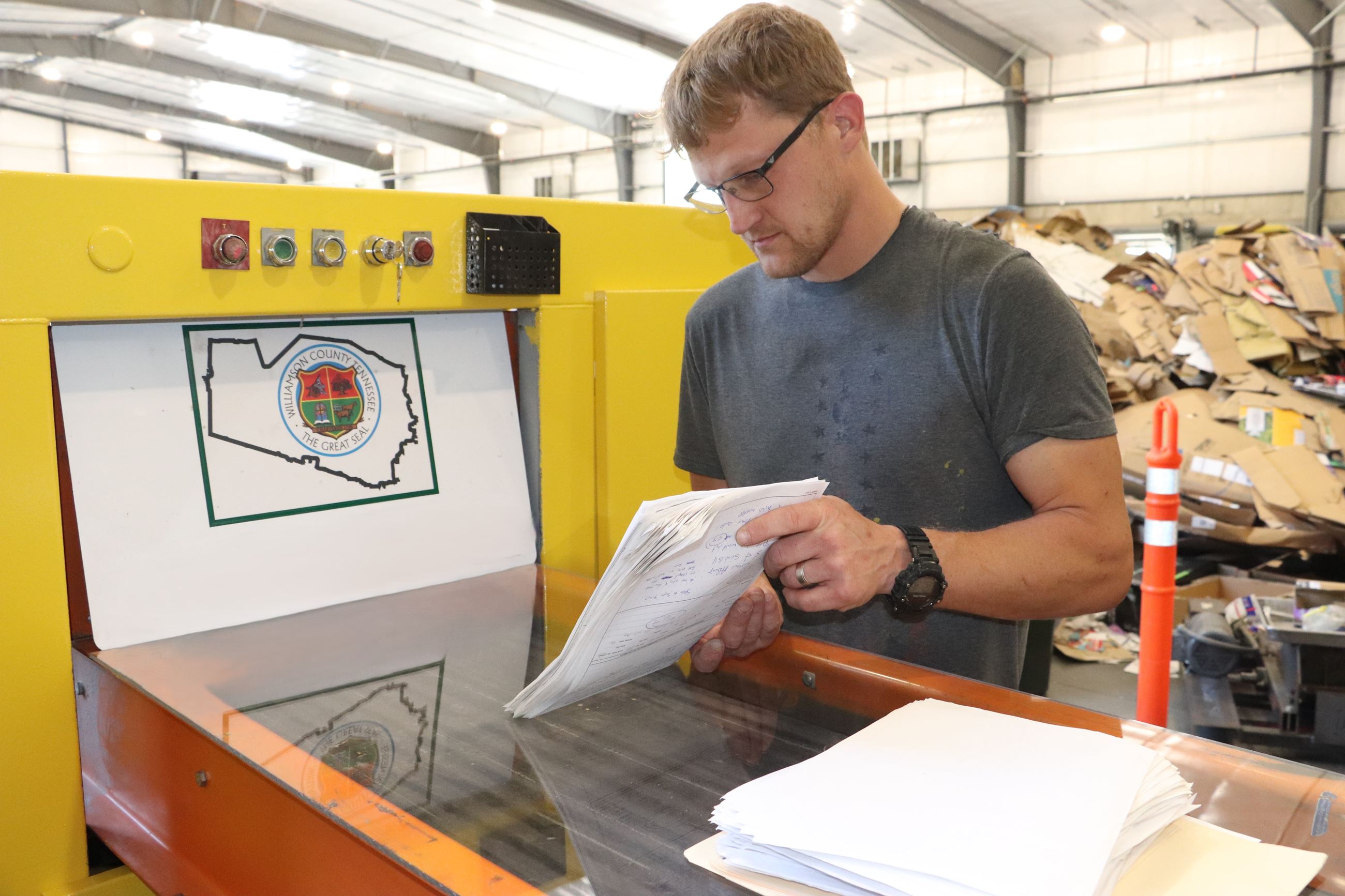 An adult feeding papers into an yellow and orange industrial paper shredder inside a warehouse